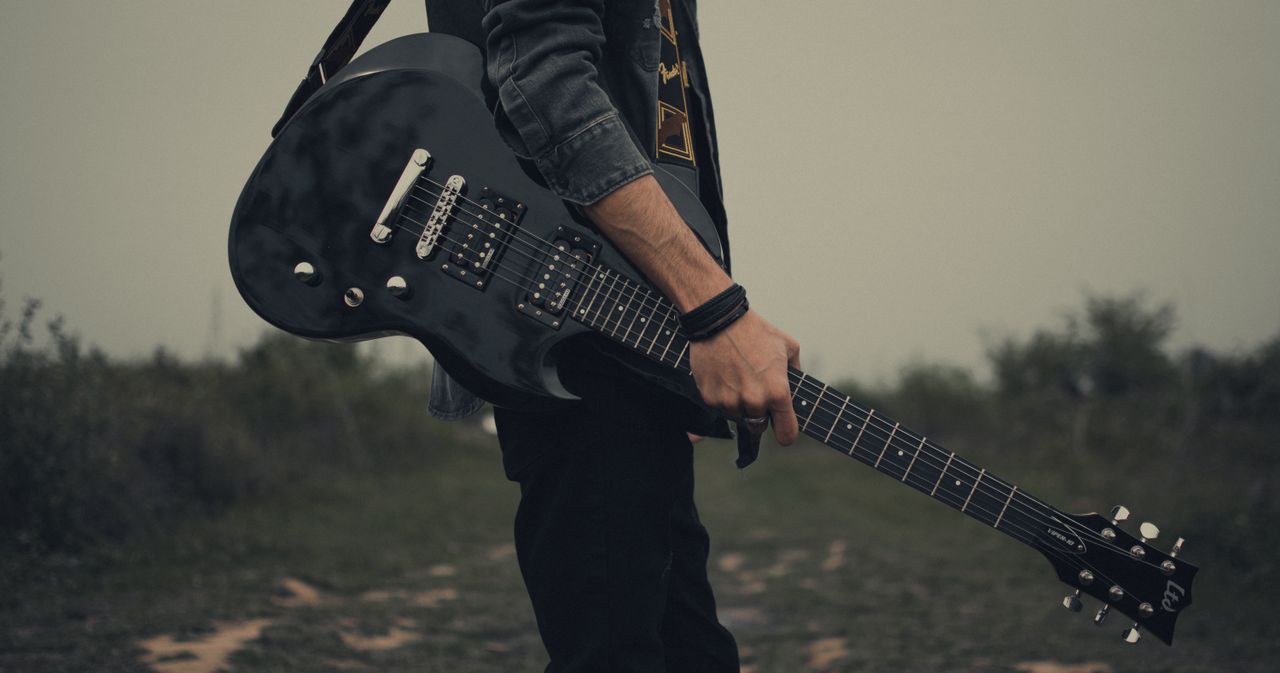 A young man holding a black electric guitar standing in an open field. Musician, outdoor rock style.